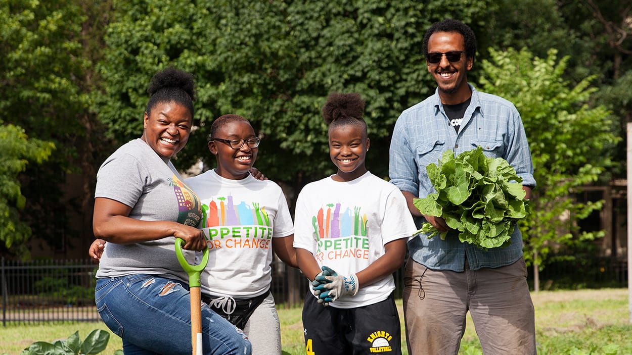 A group of adults and children pose with vegetables and garden tools.